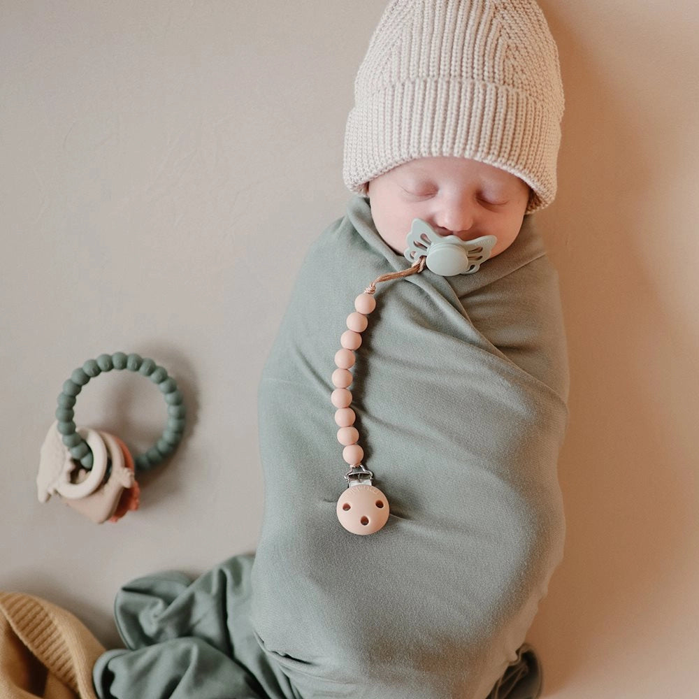 Sovende baby med FRIGG Butterfly sut samt Mushie Suttekæde og Mushie bidering Dino ved siden af